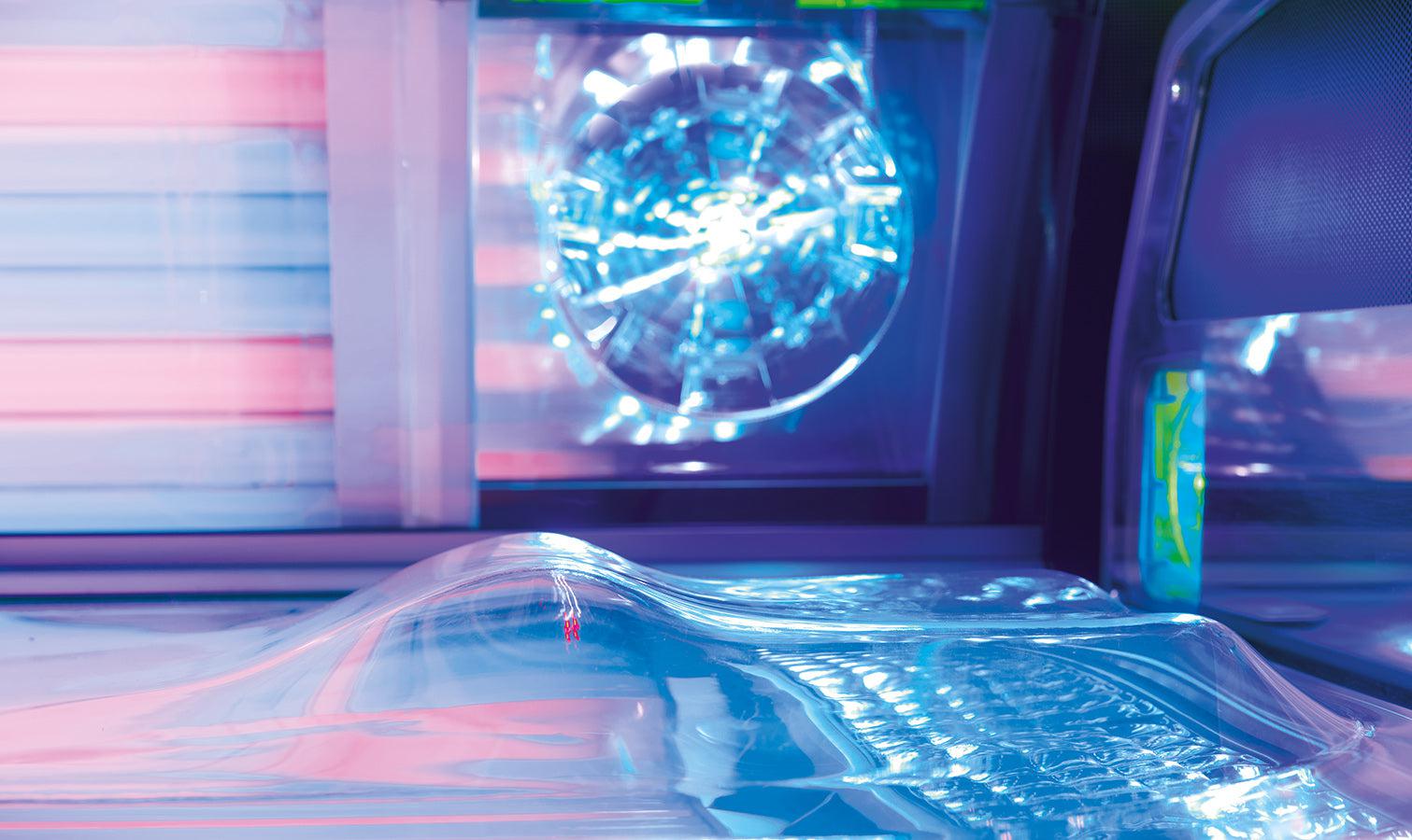 Close-up inside the Luxura tanning bed showing the contoured acrylic bench, blue and pink ambient lighting, and a bright shoulder tanner positioned in the canopy.