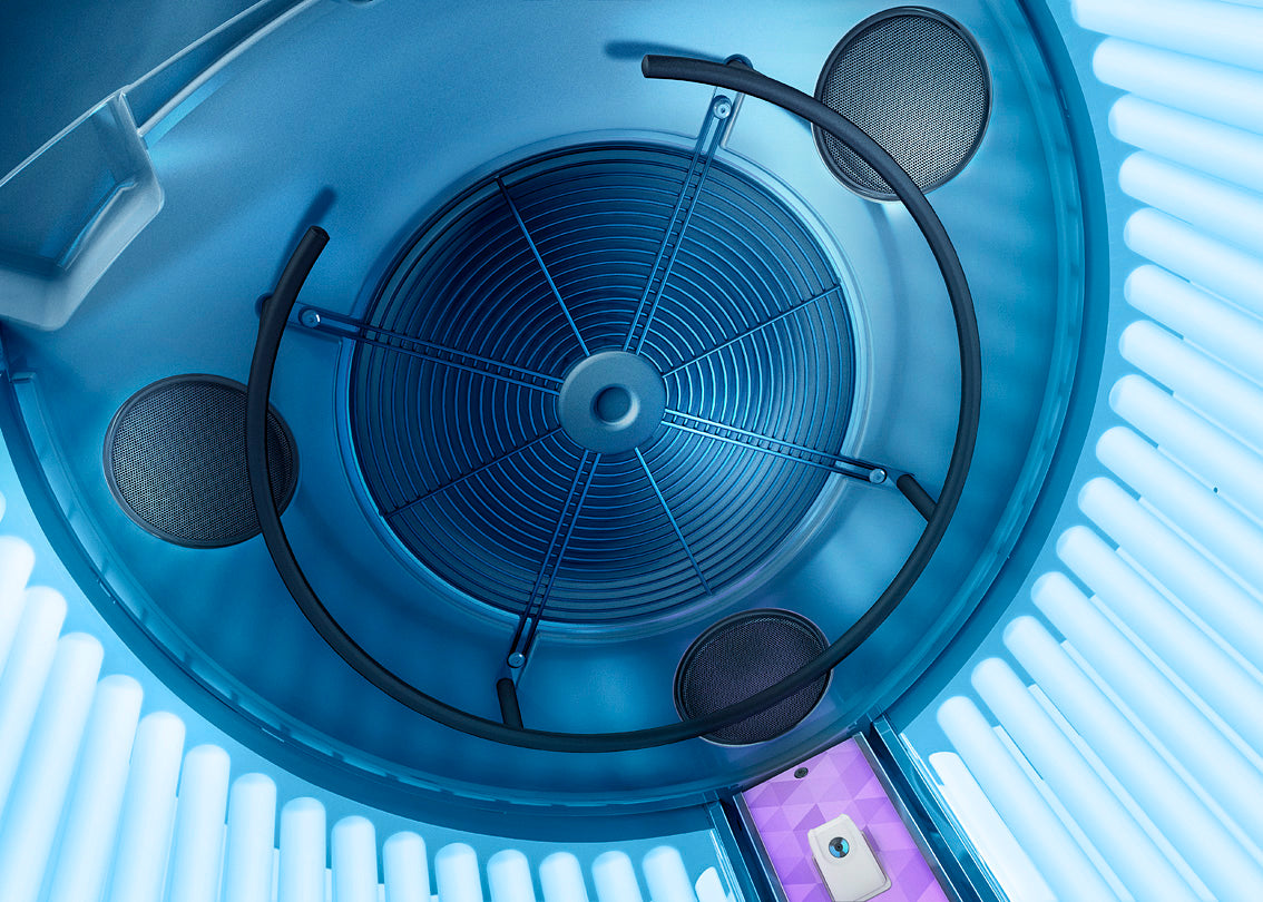 Interior ceiling view of the Luxura V8 sunbed featuring the large ventilation fan, grip bars, and built-in speakers surrounded by blue tanning lamps.